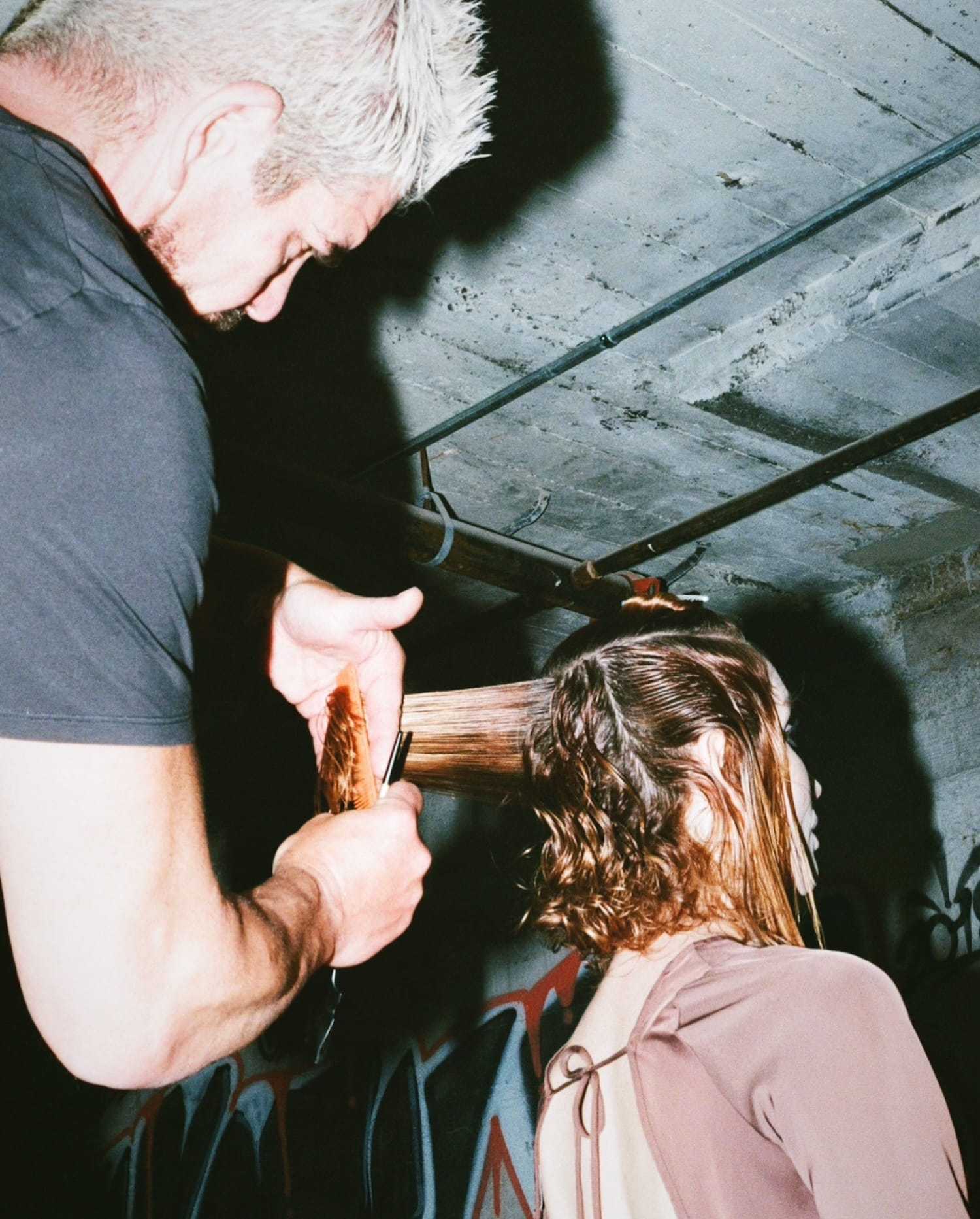 Hairstylist trims a woman's hair in an industrial space with graffiti on the walls.