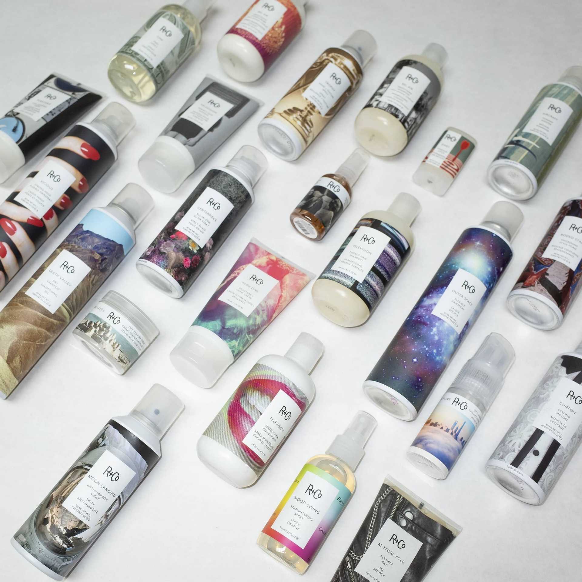 Assorted R+Co hair care products with colorful packaging arranged on a white background.