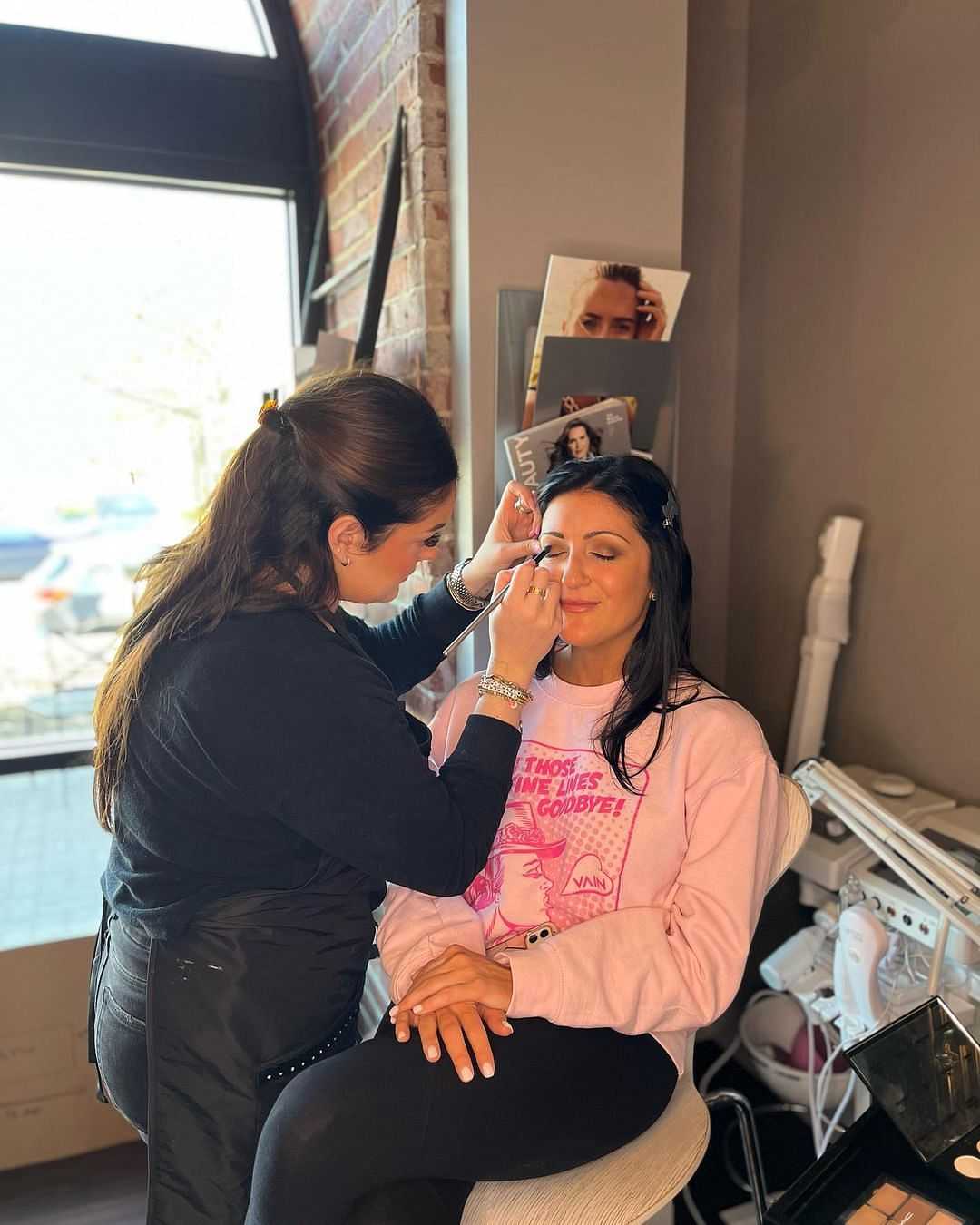 Makeup artist applying eyeshadow to a smiling woman seated in a salon chair.