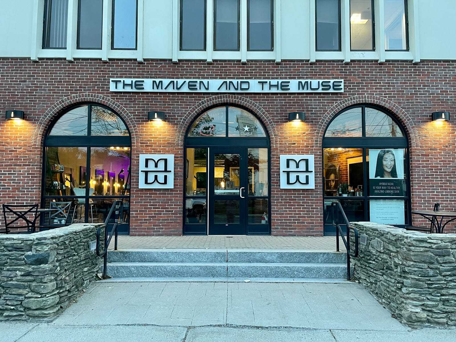 Storefront of "The Maven and The Muse" with illuminated signs and large windows on a brick building.