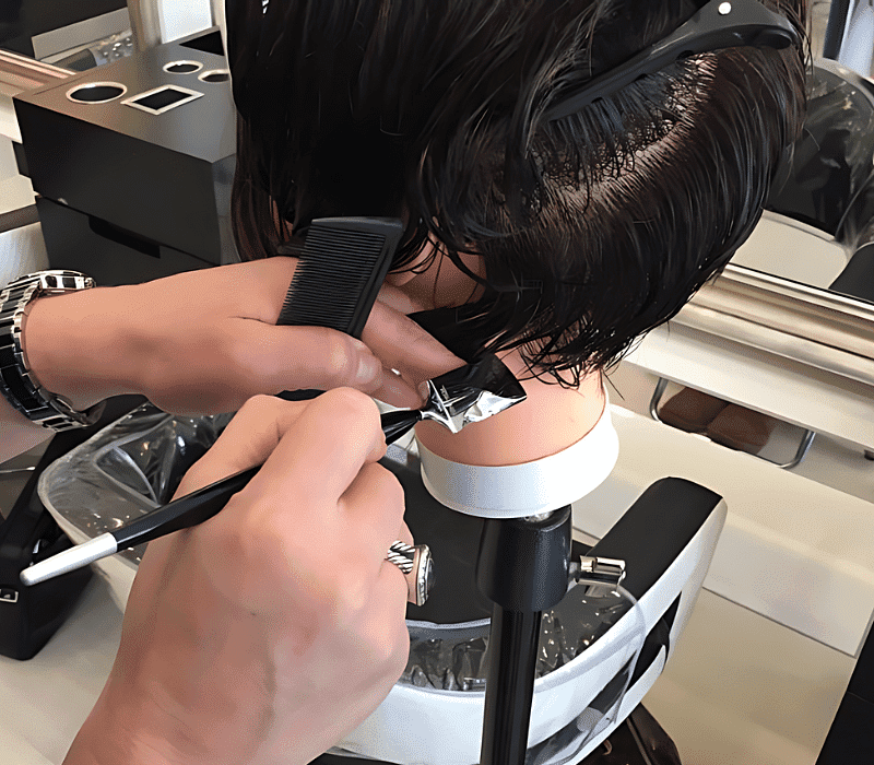 Hairdresser demonstrating cutting technique on a mannequin head in salon setting.